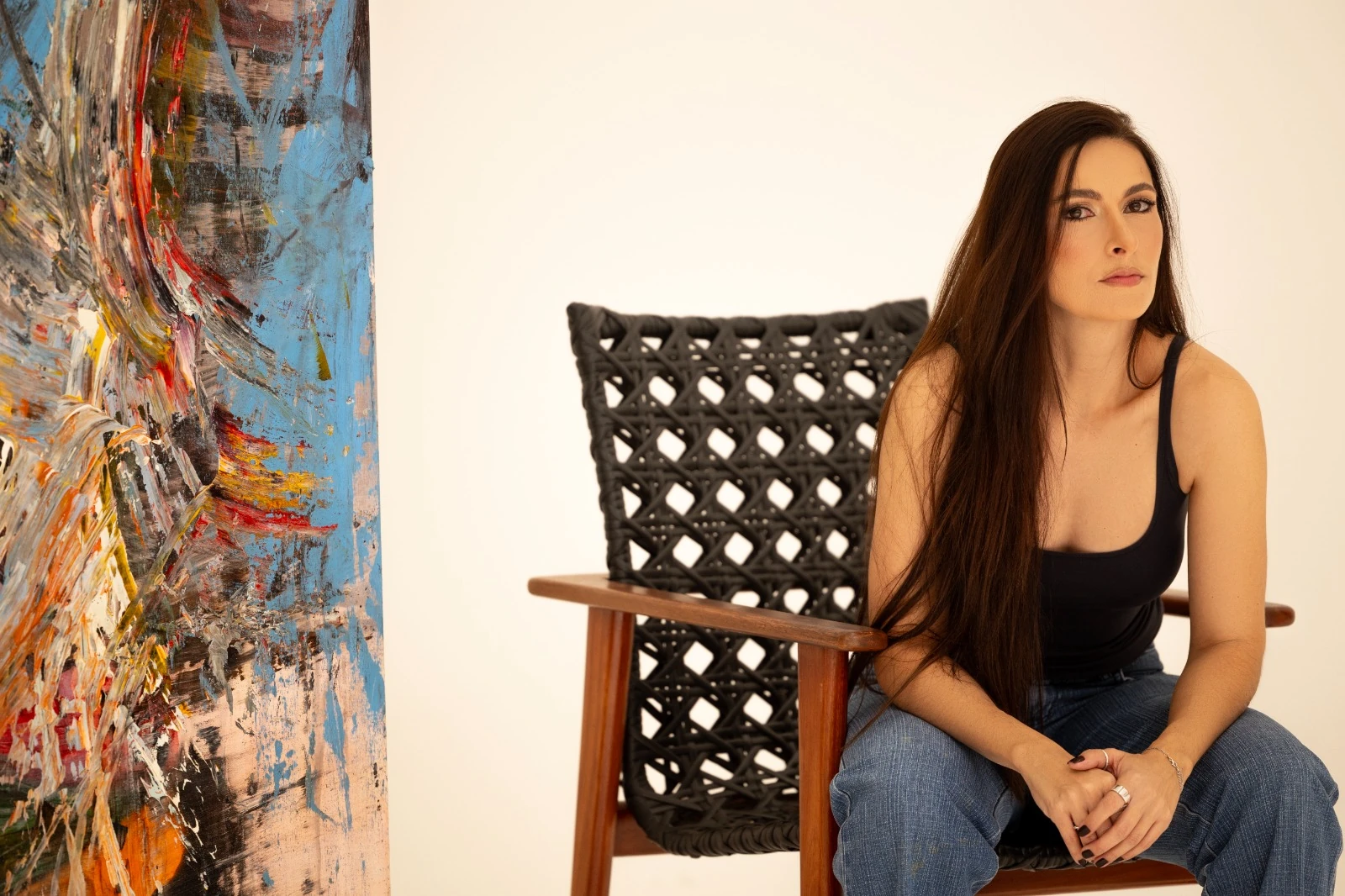 Chyara Feer sentada em uma cadeira ao lado de uma de suas pinturas.