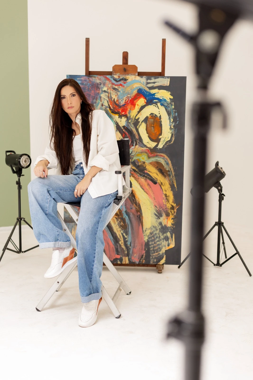Chyara Feer sentada em uma cadeira de estúdio, com uma de suas pinturas ao fundo.